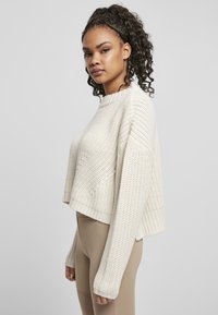 Pull en laine crocheté couleur crème avec un col large, une coupe cropped et des manches longues ; présente des poignets côtelés et un tissu texturé.