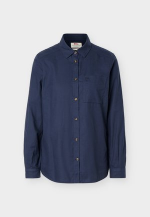 Dlhá tmavomodrá košeľa na gombíky s golierom, náprsným vreckom a béžovými gombíkmi, zobrazená na jednoduchom pozadí.