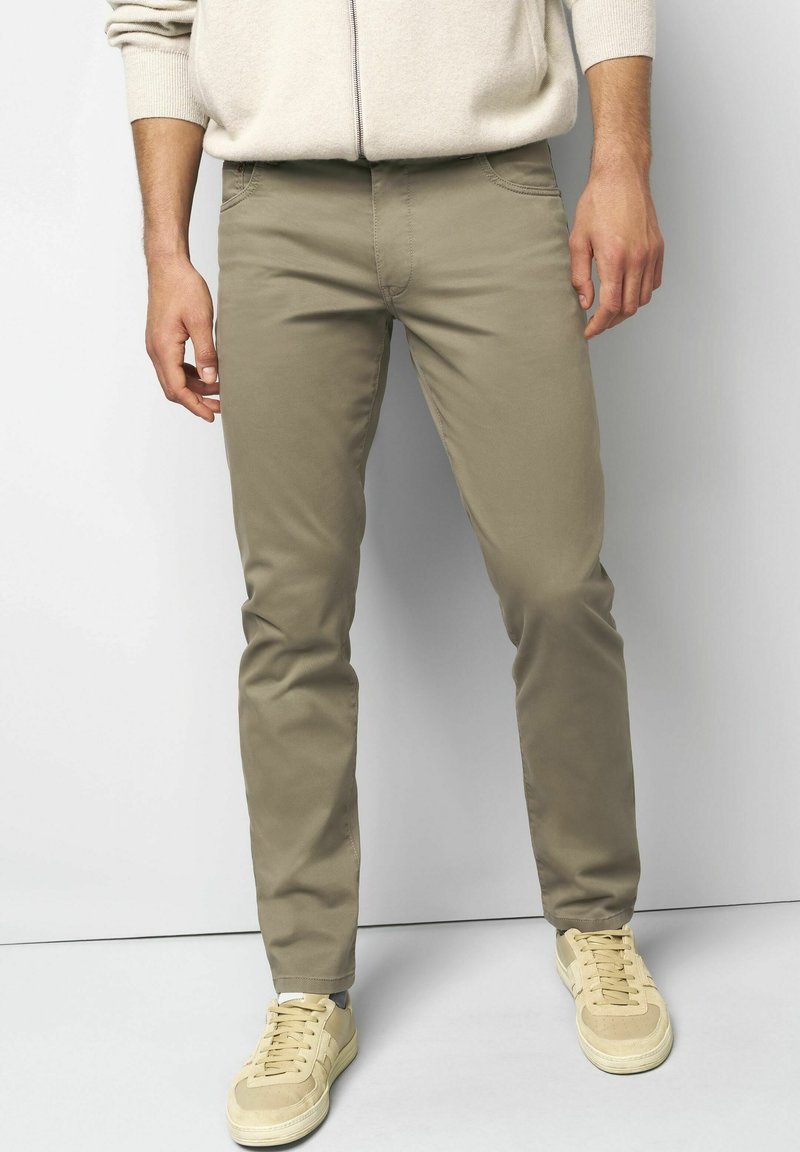 Man in beigekleurige slim-fit broek met lichte beige sneakers en een crèmekleurige sweater met rits, staand voor een witte achtergrond.