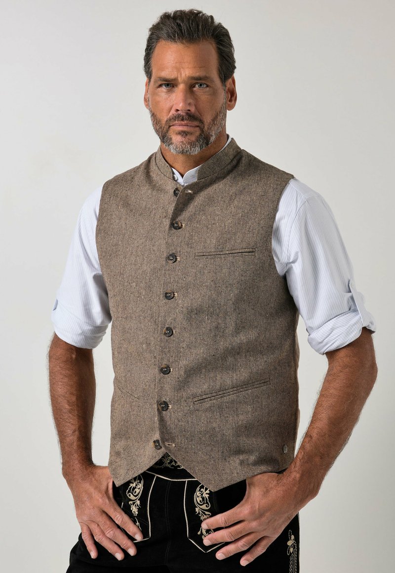 Hombre con barba corta que lleva un chaleco marrón abotonado sobre una camisa blanca remangada, de pie con las manos en las caderas contra un fondo liso.