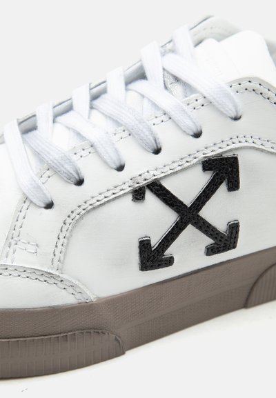 Λευκό αθλητικό παπούτσι με λευκά κορδόνια, μαύρο κεντητό λογότυπο σε σχήμα σταυρού με βέλος στη πλευρά και γκρι υφασμένο λάστιχο στη σόλα.