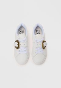 White sneakers made of smooth leather with a classic shape, featuring a decorative gold buckle and white laces on a textured sole.