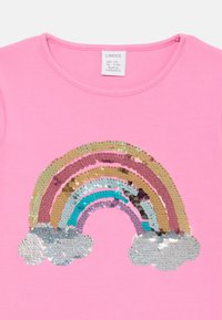 T-shirt rosa in cotone con un design di arcobaleno in paillettes nei toni dell'oro, turchese e argento, con accenti di nuvole argento sotto l'arcobaleno.