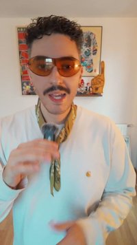 Cilvēks, kurš nēsā dzintara toņu saulesbrilles un baltu garām piedurknēm kreklu ar zelta logo, tur mikrofonu. Ap kaklu apsiets zaļš rakstains šalle.