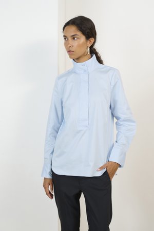 Chemise bleu clair à boutonnière en tissu lisse, avec un col haut, une patte de boutonnage et des manches longues, associée à un pantalon sombre.