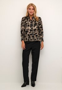 Halenka s dlouhým rukávem v abstraktním černobéžovém vzoru, vyrobena z lehkého materiálu, s zapínáním na knoflíky, v kombinaci s elegantními černými kalhotami a kotníkovými botami.