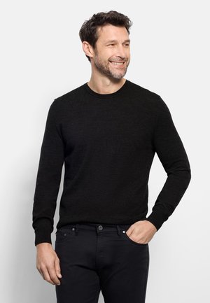 Uomo con capelli scuri e barba, indossa un maglione nero a maniche lunghe e pantaloni neri, sorride e guarda verso destra con una mano in tasca.