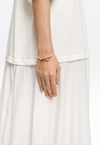 Gold heart-shaped clasp bracelet with white beaded accents, worn on a hand against a light fabric background.