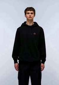 Jovem de pé, de frente, usando um hoodie preto com um pequeno logótipo circular no peito e calças pretas, contra um fundo simples.