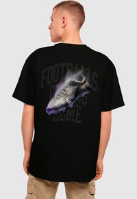 Black t-shirt featuring a graphic of a football boot with grey accents and purple smoke effects, with the text "FOOTBALL'S COMING HOME" in faded lettering.