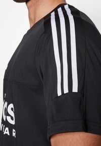 Černé sportovní tričko s bílým designem tří pruhů na rameni, s texturovanou tkaninou a výrazným bílým logem na přední straně.