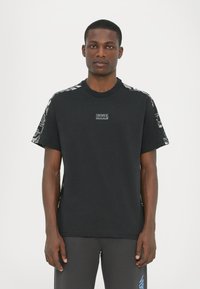 Μαύρο βαμβακερό μπλουζάκι με κοντά μανίκια, featuring υφασμάτινη διακόσμηση στους ώμους και μια μικρή ετικέτα με λογότυπο στο στήθος.