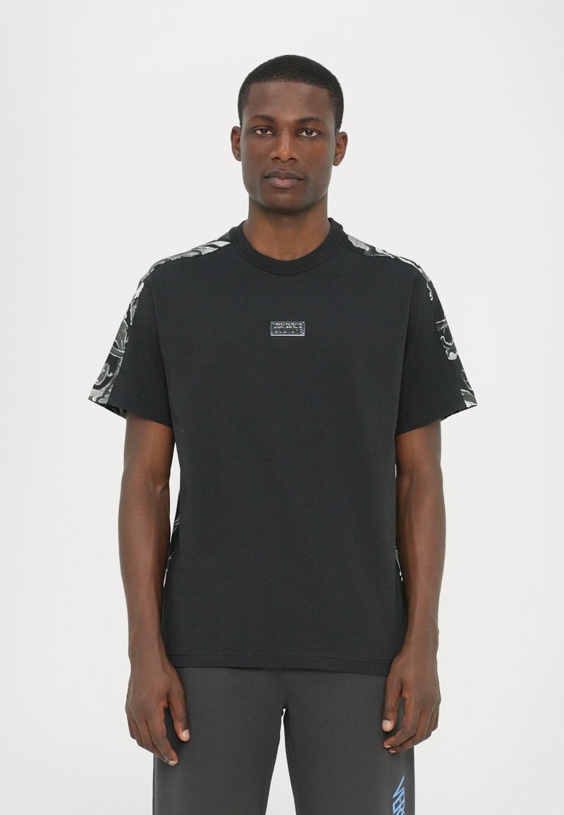 Μαύρο βαμβακερό μπλουζάκι με κοντά μανίκια, featuring υφασμάτινη διακόσμηση στους ώμους και μια μικρή ετικέτα με λογότυπο στο στήθος.