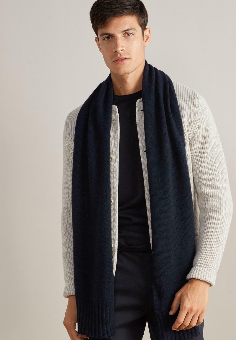 Granatowy wełniany szalik otulony wokół szyi mężczyzny, w zestawieniu z jasnoszarym prążkowanym kardiganem i ciemną koszulą.