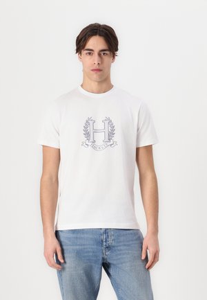 T-shirt en coton blanc avec un logo gris comportant un "H" et des détails de laurier. Manches courtes et col rond, associé à un jeans en denim bleu.
