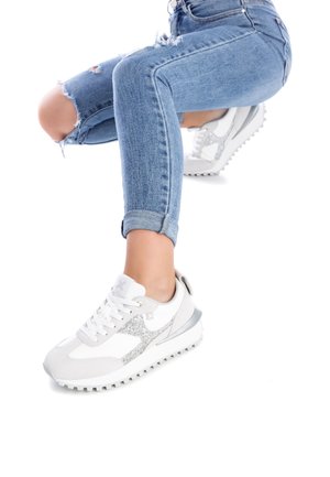 Persona che indossa jeans blu strappati e sneakers bianche con dettagli glitter argentati, accovacciata su uno sfondo bianco.