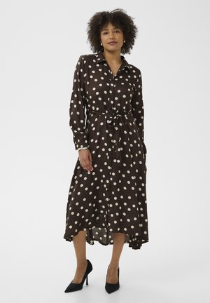 Femme portant une robe marron à manches longues boutonnée avec des pois blancs, nouée à la taille, et des talons hauts noirs, debout devant un fond blanc.