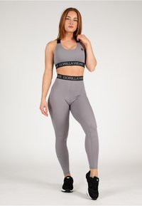 Pilkas sportinis trumpas viršus ir aukštos juosmens leggingai su juodu juostos logotipu. Modelis vilki juodus sportinius batus ir stovi prieš neutralų foną.