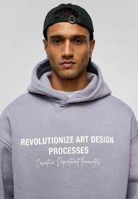Lichtpaarse hoodie gemaakt van zachte stof met grote witte tekst: "REVOLUTIONEREN ART DESIGN PROCESSES" en een artistieke slogan daaronder.