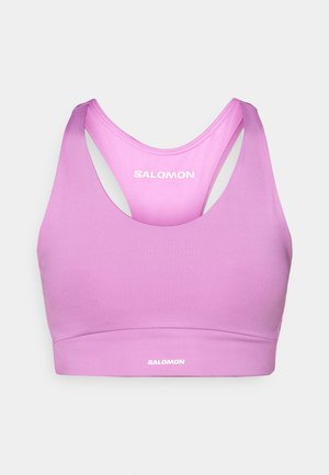 Brassière de sport rose à texture lisse, avec un col arrondi, un dos nageur et un logo. Le tissu semble extensible et respirant.