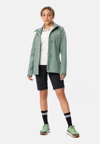 Giacca impermeabile verde chiaro con cappuccio regolabile, maglietta grigia, pantaloni corti neri e sneakers verdi con calzini a righe bianche e nere.