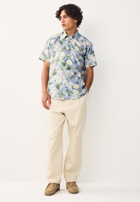 Kortærmet skjorte i lyseblå med blomsterprint; kombineret med beige bukser og brune ruskindsko. Stoffet virker blødt og let.