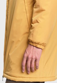 Main d'une personne portant une veste jaune avec un poignet élastique, reposant à côté de sa hanche sur un fond uni.