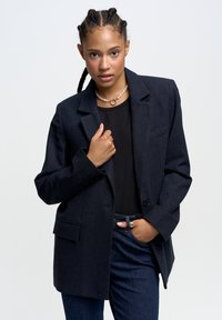 Laivastonsininen pinstripe-takki, jossa on yksi nappi, viistot kaulukset ja etutaskut, yhdistetty mustan topin ja tummansinisien farkkujen kanssa.
