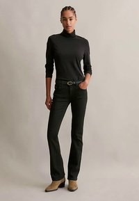 Γυναίκα με πλεγμένα μαλλιά, ντυμένη με μαύρο πουλόβερ, μαύρα τζιν με ζώνη και μπεζ μποτάκια μέχρι τον αστράγαλο, που στέκεται μπροστά σε απλή φόντο.