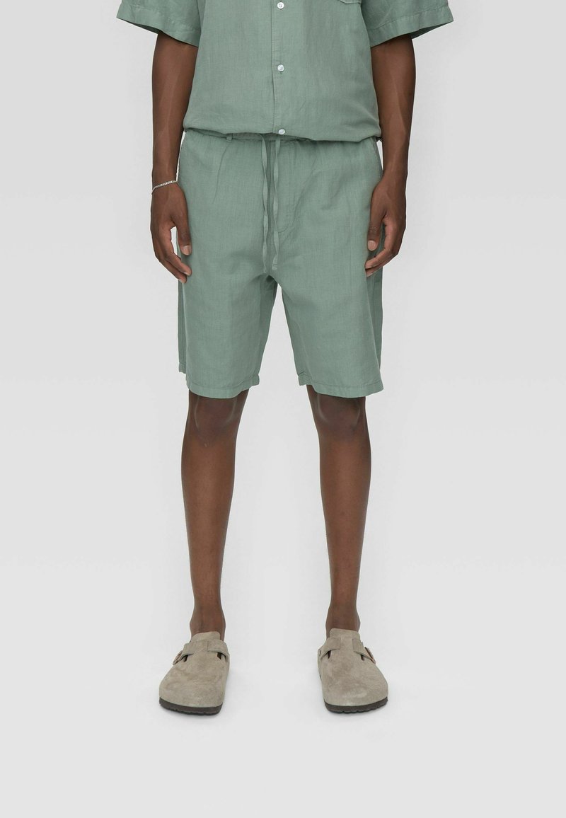 Ensemble chemise en lin vert clair et shorts avec une coupe décontractée et une taille à cordon de serrage, associé à des chaussures beiges à enfiler.