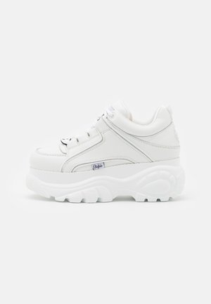 Biały sneaker na platformie z grubą podeszwą, zapinany na sznurowadła z przodu oraz małą oznakowaną metką po boku, pokazany z boku na jednolitym tle.