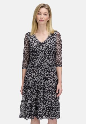 Femme aux cheveux blonds portant une robe imprimée léopard noir et gris, longueur genou, avec des manches trois-quarts transparentes, se tenant devant un fond blanc.
