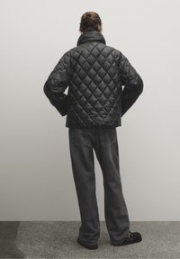 Czarna pikowana kurtka puchowa z wysokim kołnierzem, z długimi rękawami i pudełkowatym krojem, zestawiona z ciemnymi dżinsami i czarnym obuwiem.