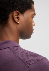 Giovane uomo con i capelli ricci corti che indossa una maglietta a maglia testurizzata viola, mostrato di lato e di schiena su uno sfondo grigio uniforme.