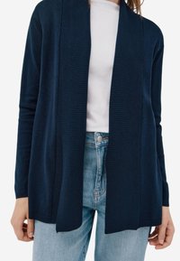 Cardigan marine à design ouvert à rayures. Fabriqué dans un matériau doux, il présente des manches longues et une coupe décontractée, associé à un jean clair.