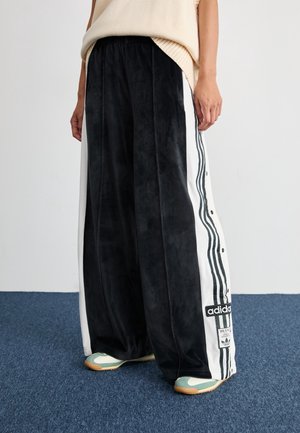 Personne portant un pantalon de survêtement large noir Adidas avec des bandes blanches sur les côtés et des boutons, debout sur un tapis bleu avec des baskets vertes et blanches.