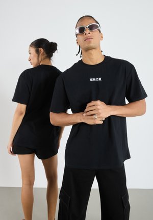 Sort t-shirt med korte ærmer lavet af bomuld, med hvid tekst på brystet. Modellen parrer den med mørke shorts og cargo-bukser.