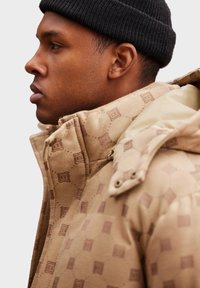 Béžová vzorovaná péřová bunda s vysokým límcem a hladkým podšíváním, s jemnými embosovanými detaily. V kombinaci s černou pletenou čepicí.