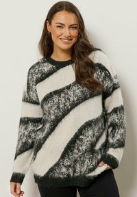 Frau mit langen braunen Haaren, die einen schwarz-weißen, abstrakt gemusterten Pullover trägt, lächelt mit einer Hand in der Tasche vor einem schlichten Hintergrund.