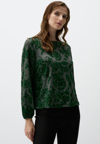 Camicia a maniche lunghe con motivo floreale verde, in un tessuto morbido, caratterizzata da una vestibilità comoda e polsini elasticizzati, abbinata a pantaloni neri.