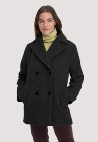 Dunkelgrauer, zweireihiger Wollmantel mit großen schwarzen Knöpfen und einem Reverskragen; kombiniert mit einem hellgrünen Rollkragenpullover darunter.