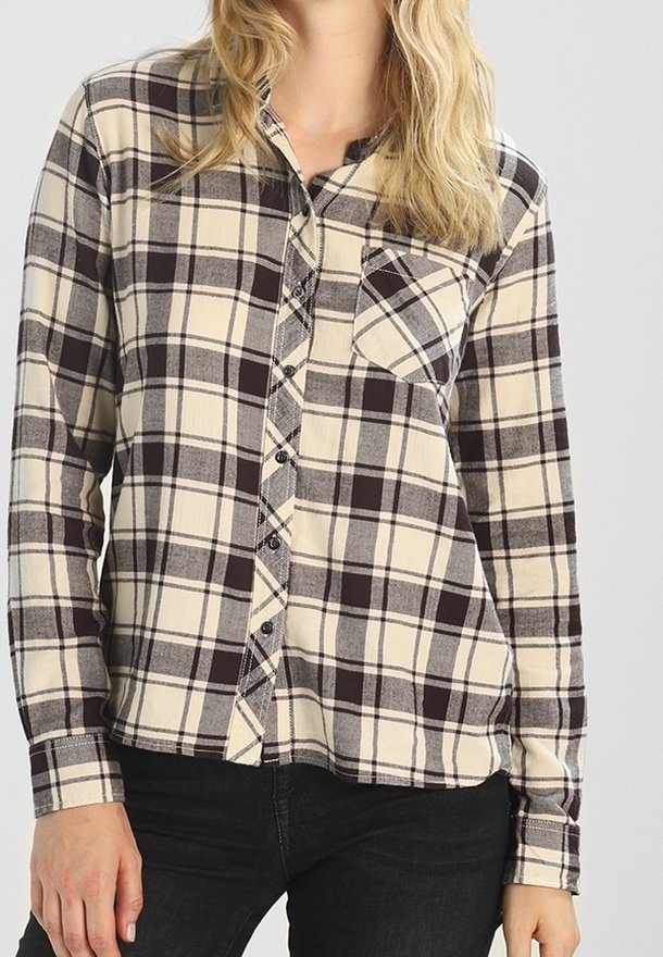 Femme portant une chemise à carreaux beige et noire boutonnée avec une poche poitrine et des manches longues, associée à un pantalon noir.