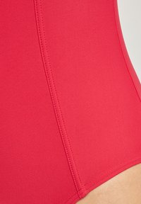 Close-up van de rode badpakstof, met een gladde textuur, schone stiksels en een aansluitende vorm. Geen patronen of extra accenten zichtbaar.