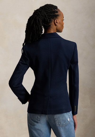 Γυναίκα με μακριά κοτσίδια, φορώντας μπλε σκούρο σακάκι και μπλε τζιν, στέκεται με την πλάτη σε ουδέτερο φόντο.