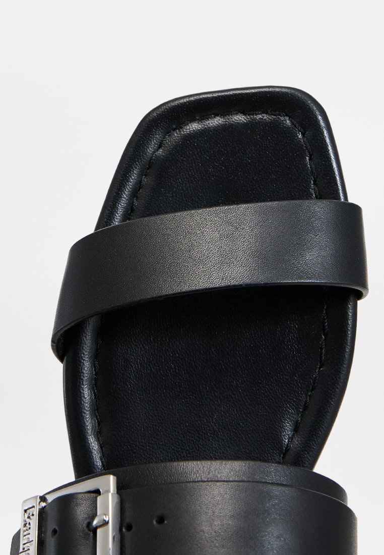 Czarna skórzana sandał z szerokim paskiem na stopie, płaską podeszwą i błyszczącym srebrnym zapięciem. Gładka struktura i minimalistyczny design.