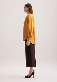 Senfgelbe Bluse mit langen, puffartigen Ärmeln, kombiniert mit einem taillierten, knielangen braunen Rock und schwarzen Stilettos.