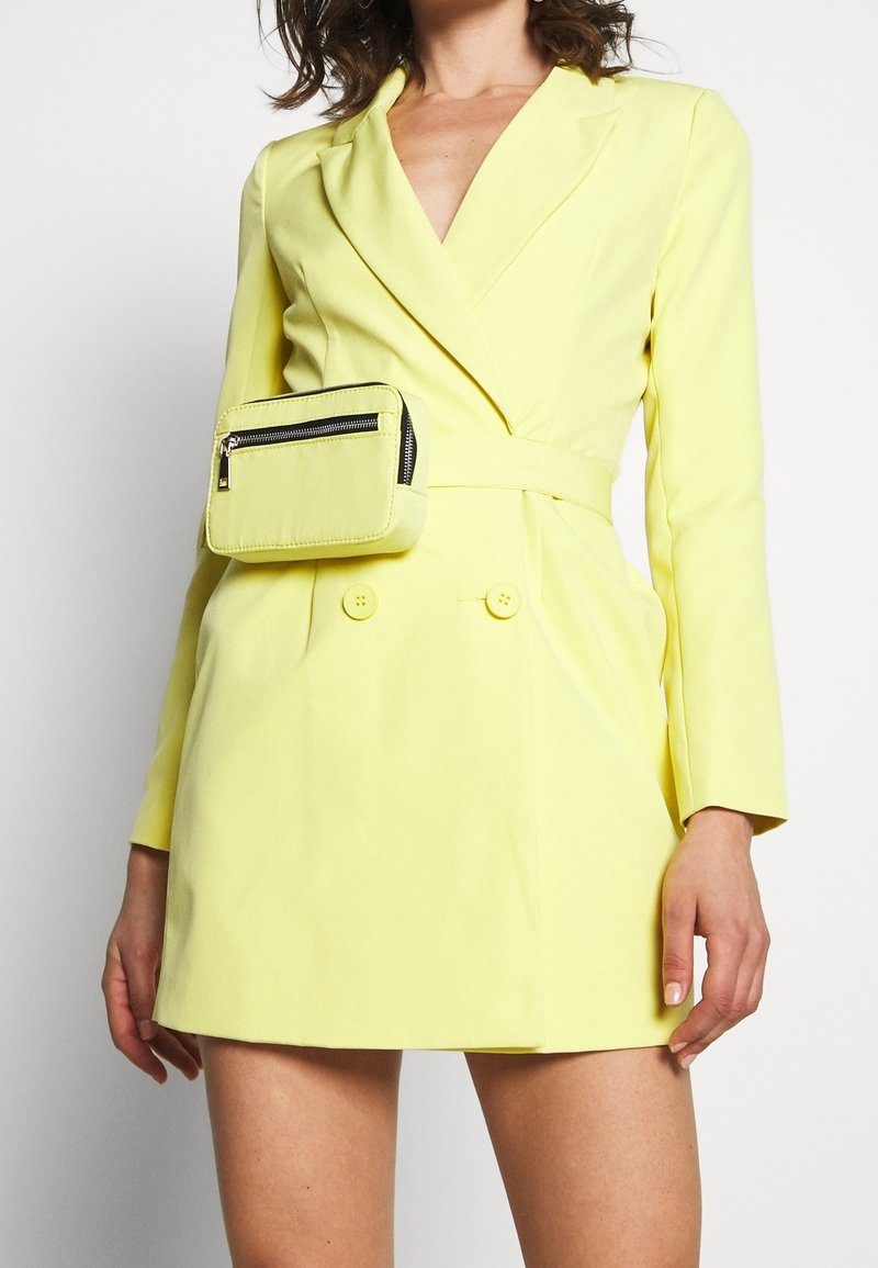 Femme portant une robe blazer jaune vif avec des boutons doubles et un sac ceinture jaune assorti zippé à la taille.