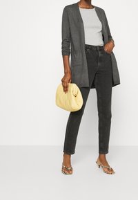 Kvinna som bär en grå kofta, ljusgrå topp, svarta jeans, hälskor med ormskinnsmönster och håller en mjuk gul clutch-väska.