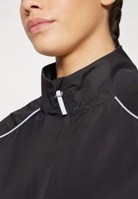 Zwarte windbreakerjas met een hoge kraag, met grijze accenten en een rits aan de voorkant. De stof lijkt glad en lichtgewicht.