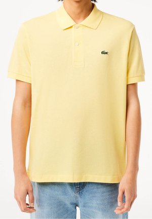 Personne portant un polo à manches courtes jaune clair avec un petit logo de crocodile vert sur le côté gauche de la poitrine, associé à un jean bleu clair.